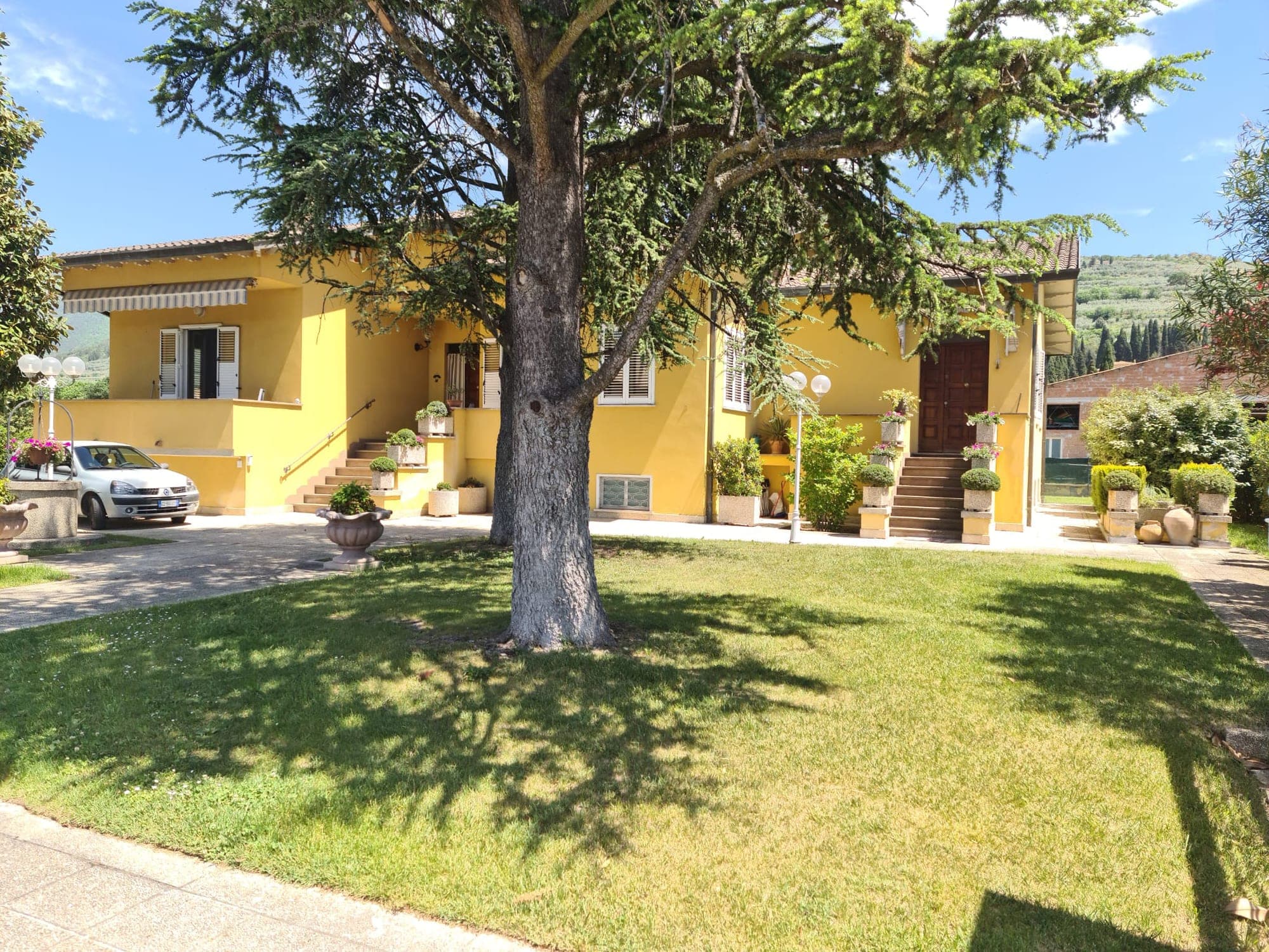 La villa gialla immersa nel verde, con il grande cedro e le colline di Spello